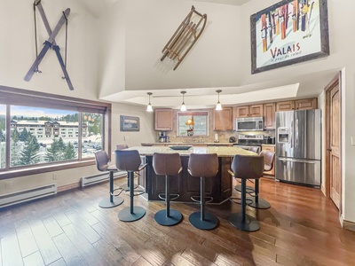 Modern kitchen with wooden floors, a central island with five stools, stainless steel appliances, and ski-themed decor adorning the walls. Large window offers a view of a building and trees outside.