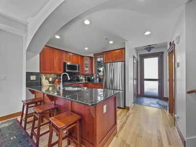 A modern kitchen with dark granite countertops, wooden cabinets, stainless steel appliances, and a wooden island with two stools. The room has recessed lighting and a tiled floor near the back door.