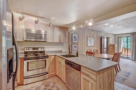 A modern kitchen with stainless steel appliances, light wood cabinets, and a granite countertop island. Adjoining dining area with a wooden table and chairs, and a view of a balcony with outdoor greenery.