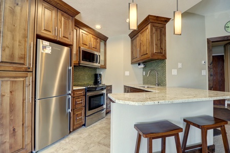 A modern kitchen with wooden cabinets, stainless steel appliances, a tiled backsplash, and a granite countertop bar with two wooden stools. Two pendant lights hang from the ceiling.