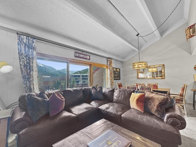 A living room with a large brown sectional sofa, patterned pillows, a large window with a scenic mountain view, a dining area in the background, and modern light fixtures.