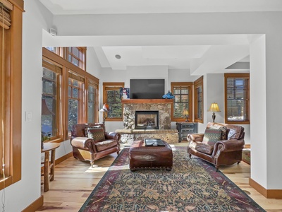 A cozy living room with leather armchairs, a large rug, wooden floors, multiple windows, and a stone fireplace with a TV above it.