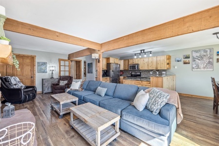 An open-concept living room and kitchen area featuring a blue sectional sofa, wooden furniture, and a mix of modern and rustic decor. The kitchen has stainless steel appliances and wooden cabinetry.