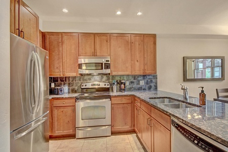 A modern kitchen with stainless steel appliances, wooden cabinetry, a granite countertop, and a double-basin sink. A mirrored wall accent is visible on the right.