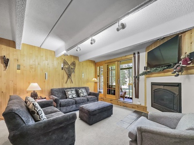 A cozy living room features grey upholstered furniture, a fireplace, wood-paneled walls with decorative items, and large windows leading to a sunlit room.