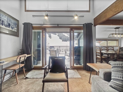 A cozy living room with large windows, a balcony view of snow-covered mountains, a wooden dining table with chairs, a desk with a chair, and a sofa beside a wooden coffee table.