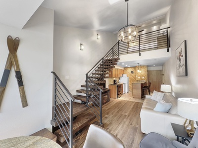 Living area with a staircase leading to a loft, wooden flooring, modern kitchen, and a leather sofa. Wall-mounted skis for decoration, and a hanging light fixture.