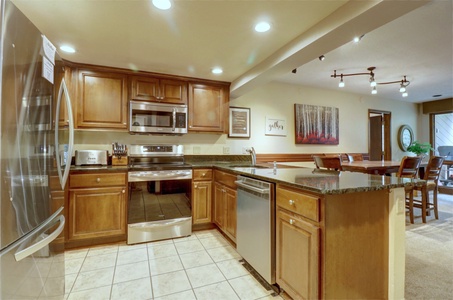 A modern kitchen with stainless steel appliances, wooden cabinets, granite countertops, and a tile floor. The kitchen opens to a dining area with a wooden table, chairs, and artwork on the wall.