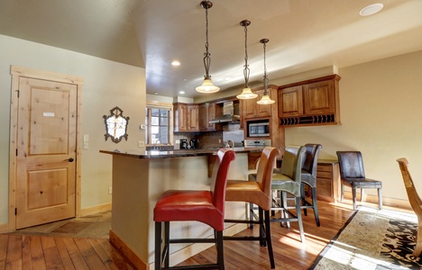 A modern kitchen with wooden cabinets and a central island with colorful bar stools. Three pendant lights hang above the island, and the room includes a microwave, stove, and wooden door.