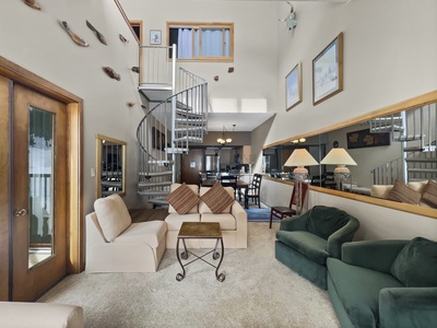 A living room with beige and green seating, a metal spiral staircase, large mirrors, and a wooden door with glass panels. The space is well-lit, featuring decor items on the walls and shelves.