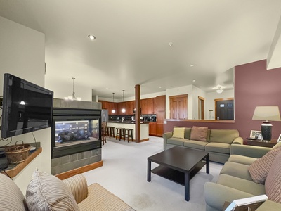 A living room with beige couches, a striped armchair, coffee table, wall-mounted TV, and a double-sided fireplace. The background shows a kitchen with bar stools at a counter.