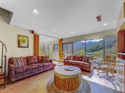 A brightly lit living room with two brown sofas, a round coffee table, a spiral staircase, large windows, and a scenic mountain view.