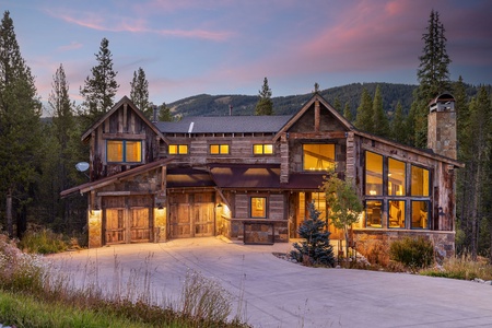 A rustic, mountain-style house with large glass windows and a wooden exterior, surrounded by trees and mountains, at dusk. The home is warmly lit from inside, with a large driveway.