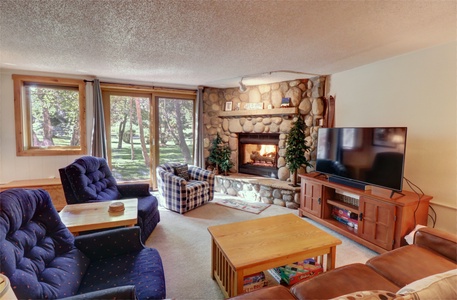 A cozy living room with a stone fireplace, TV, and comfortable seating including blue and plaid armchairs. A large window offers a view of the trees outside.