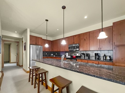 A modern kitchen featuring wooden cabinets, granite countertops, stainless steel appliances, and hanging pendant lights. A breakfast bar with three wooden stools is in the foreground.