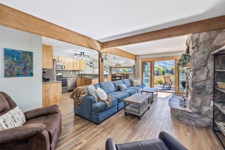 A spacious living room with wooden beams features a blue sofa, stone fireplace, large windows with mountain views, and an adjacent kitchen and dining area with modern appliances.