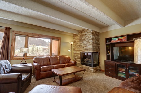 A cozy living room with a brown leather sofa and chairs, a wooden coffee table, a stone fireplace, a flat-screen TV on a wooden stand, and large windows with a mountain view.