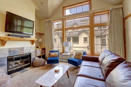 A cozy living room with a large leather couch, two blue chairs, a TV mounted above a fireplace, a coffee table, and large windows letting natural light in.