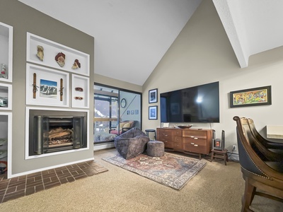 A living room with a high ceiling, a mounted TV, a fireplace, artwork on the walls, and a mix of modern and traditional furniture. The room features a mountain view from a large window.