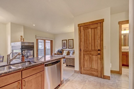 A modern kitchen with a granite countertop and stainless steel dishwasher opens to a cozy living area. The room features wooden cabinetry, a fireplace, a small dining area, and a doorway leading to a bathroom.