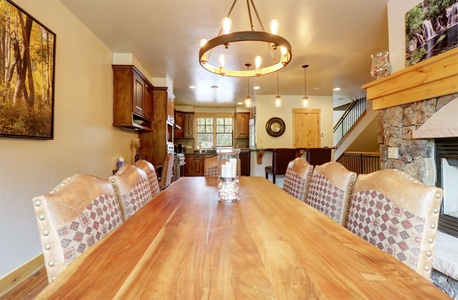 A dining room with a long wooden table, upholstered chairs, a circular chandelier, and a view of the kitchen. A stone fireplace and framed artwork are visible on the right wall.