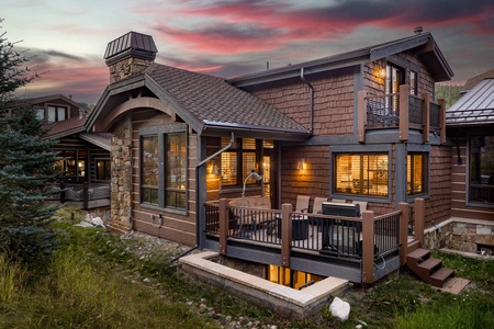 Modern rustic house with large windows, stone chimney, and wooden siding. The house features an elevated deck with outdoor furniture and warm interior lighting, set against a sunset sky.