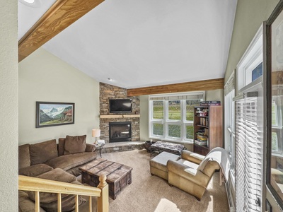 A living room with a brown sectional sofa, a stone fireplace, a wooden coffee table, and a bookshelf. The room features a large window and exposed wooden beams on a high ceiling.