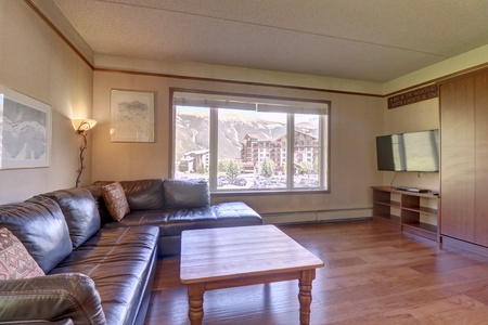 Living room with brown leather sectional sofa, wooden coffee table, wall-mounted flat-screen TV, and a large window with a mountain resort view. Wooden flooring and minimalistic decor.