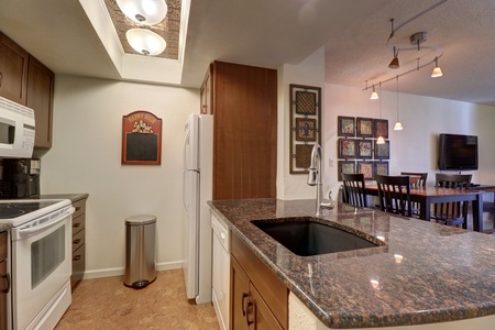 Modern kitchen with brown cabinets, granite countertops, stainless steel sink, white appliances, and adjacent dining area with black table and chairs. Wall art decorates the dining space.