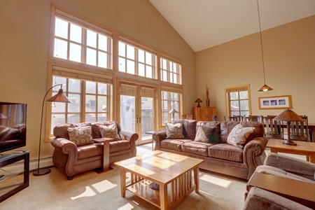 Living room with large windows, high ceiling, brown leather sofas, a wooden coffee table, two side tables, lamps, and a flat-screen TV. Warm sunlight fills the space.