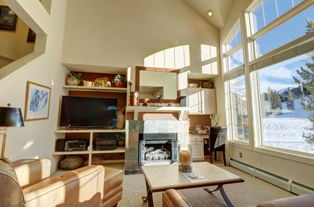 A living room with high ceilings, large windows, a fireplace, and a wall-mounted TV. The room has a view of East Village ski slopes at Copper Mountain.