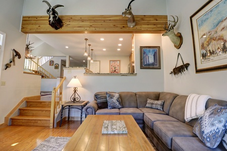 Living room with gray sectional sofa, wooden coffee table, mounted animal heads on walls, and visible staircase leading to the upper floor. The room is well-lit with ceiling lights and a floor lamp.