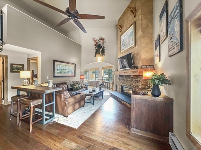 A spacious living room with a high ceiling features a fireplace, leather couch, wooden floors, and large windows. Mounted artworks and decorations adorn the walls, and a ceiling fan hangs overhead.