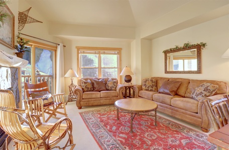 A cozy living room with two brown sofas, a round wooden coffee table, and rustic rocking chairs. The room features large windows, a decorative rug, framed art, and a mirror above one sofa.