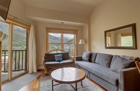 A cozy living room features a gray sofa, a round wooden coffee table, and a window with a mountain view. A balcony with a view of the same mountains can be seen through a glass door to the left.