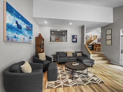 A modern living room features a gray sectional sofa, two matching chairs, a round coffee table, a geometric rug, and a painting of a bear. A staircase leads up to a dining area with a wooden table.