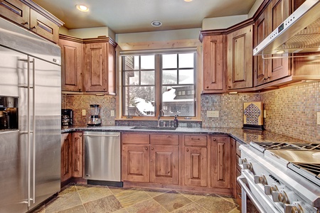 A modern kitchen with wooden cabinets, stainless steel appliances, including a refrigerator, dishwasher, and stove, and a window with a view outside. The backsplash is tiled, and the floor is stone.