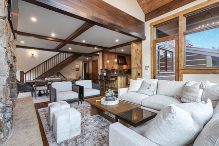 A spacious living room with a white sofa, two armchairs, a wooden coffee table, and a kitchen area in the background. The room features wooden accents and a staircase leading upstairs.