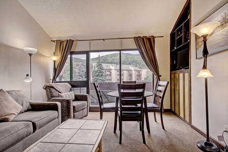 A living room with a grey sofa, armchair, coffee table, and dining table with four chairs. Floor lamps are present, and a large window with curtains offers a view of buildings and trees.