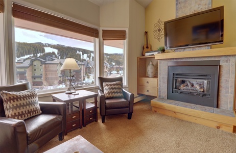 A cozy living room with two leather chairs, a fireplace, a flat screen TV, and large windows offering a mountain view.