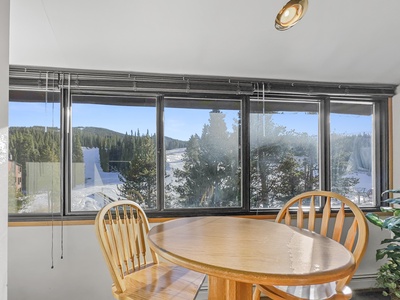 A round wooden table with three matching chairs is positioned near large windows with views of a snowy landscape and evergreen trees.