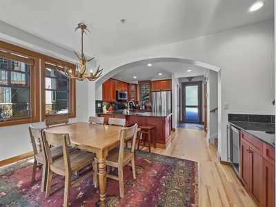 A wooden dining table with four chairs is placed on a patterned rug in an open-plan kitchen and dining area. The kitchen features stainless steel appliances, wooden cabinets, and a center island with stools.