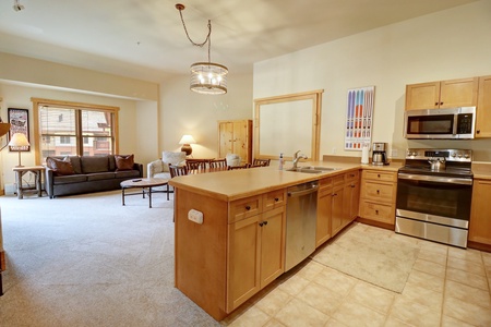 A spacious kitchen and living area featuring wooden cabinets, stainless steel appliances, a sofa, and a coffee table. A large island separates the kitchen from the living space, and light pours in from windows.