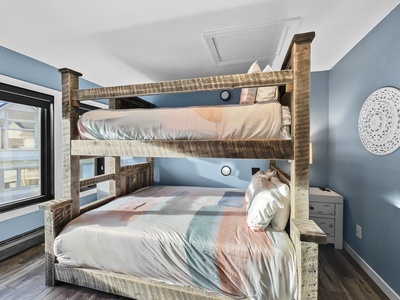 A wooden bunk bed with white and pastel-colored bedding is placed against a blue wall in a well-lit room with hardwood floors.