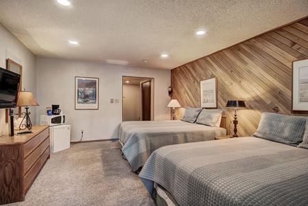 Hotel room with two beds covered in light gray bedspreads, a wooden dresser holding a TV, a small fridge, and coffee maker. Walls feature framed pictures and a chevron wooden panel. Warm lighting.