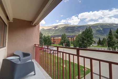 A balcony with two chairs and a small table overlooks a street, parking lot, and distant mountains under a partly cloudy sky.