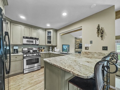 A modern kitchen with stainless steel appliances, granite countertops, light green cabinetry, and bar seating. The adjacent open area features a fireplace and a wall-mounted TV.