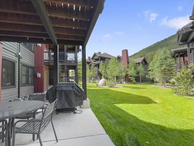 A patio with a barbecue grill and table set overlooks a grassy courtyard surrounded by multi-story residential buildings and evergreen trees on a clear day.