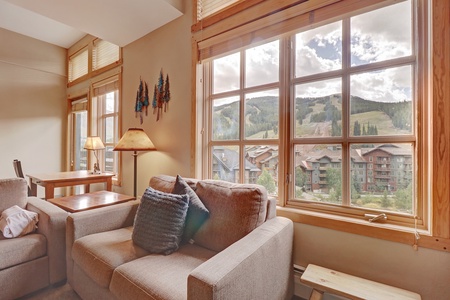 Sun-filled corner of the condo’s living area, featuring a plush armchair and rustic wood accents. Large windows frame sweeping views of the Center Village rooftops and the ski slopes beyond.