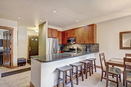 A modern kitchen with a black granite countertop, wooden cabinets, stainless steel appliances, and a row of four wooden stools at a breakfast bar. A dining area is visible to the right.